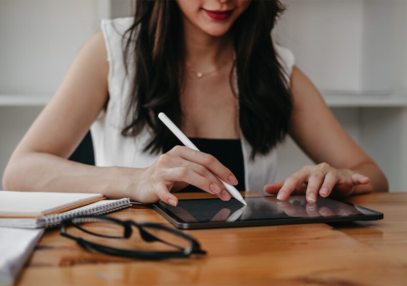 Close-up of a woman using an Apple Pencil on a digital planner with a tablet, accompanied by a notebook and glasses on a wooden desk