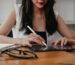 Close-up of a woman using an Apple Pencil on a digital planner with a tablet, accompanied by a notebook and glasses on a wooden desk